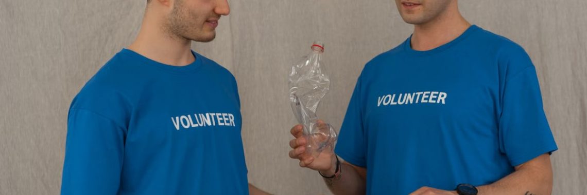 Two male volunteers in blue shirts collecting plastic bottles for recycling indoors.