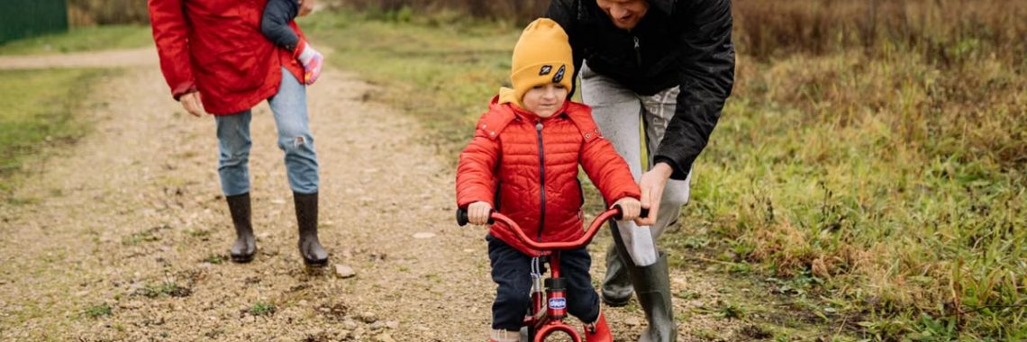Happy family walking outdoors in fall, teaching child to ride bike.